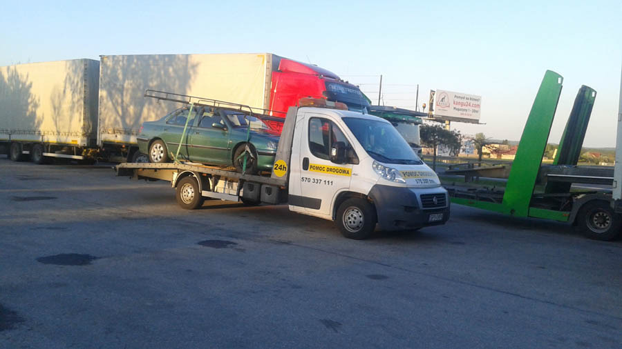 Laweta z zielonym samochodem osobowym na platformie, zaparkowana obok zestawu ciężarowego z naczepami, w tle widoczny billboard.