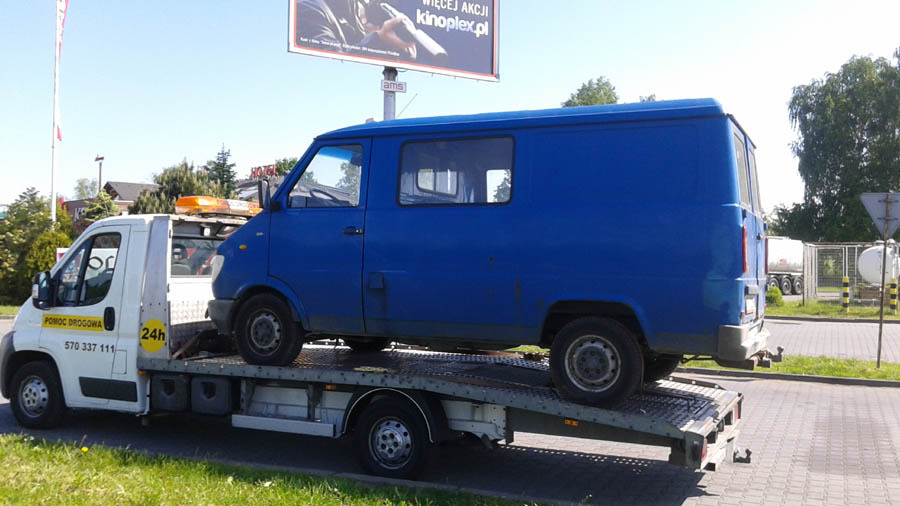 Niebieski bus na lawecie pomocy drogowej z widocznym numerem telefonu i informacją o dostępności 24h, na tle billboardu reklamowego.