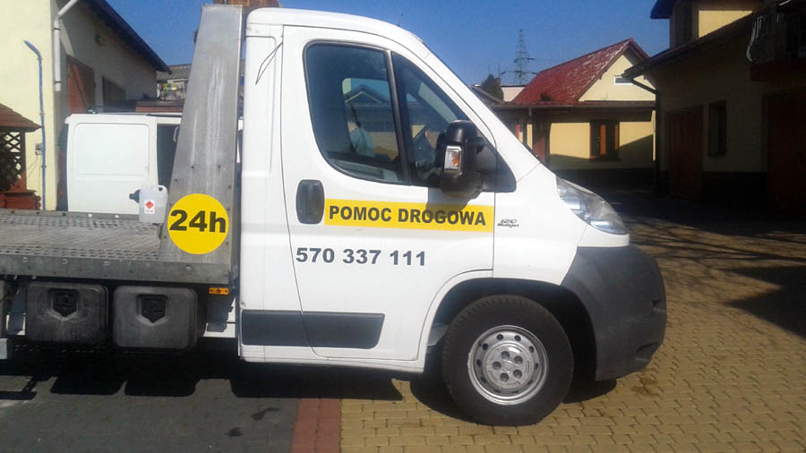 Biała laweta z napisem 'Pomoc Drogowa' i numerem telefonu, zaparkowana na brukowanym podjeździe przed budynkiem w słoneczny dzień.