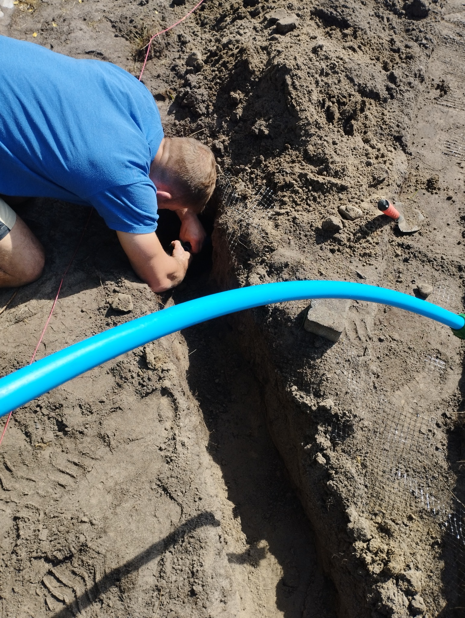 Instalacja niebieskiej rury w wykopie ziemnym, widoczny pracownik w niebieskiej koszulce, ziemia, siatka wzmacniająca i fragment czerwonego sznurka wyznaczającego trasę.