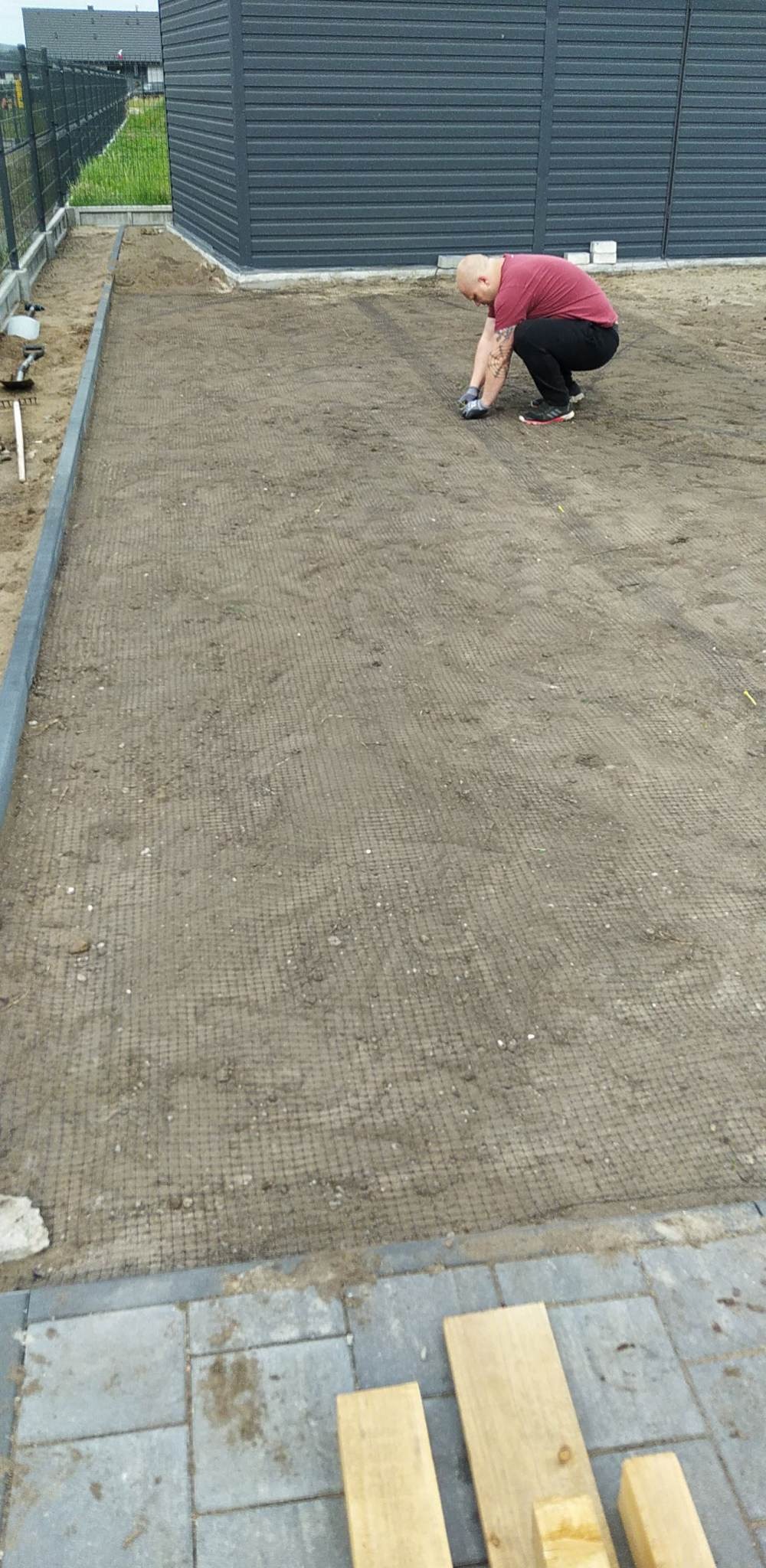 Przygotowanie podłoża pod trawnik z rolki: mężczyzna w rękawicach wyrównuje ziemię pokrytą siatką wzmacniającą, widoczne obrzeża i drewniane deski.