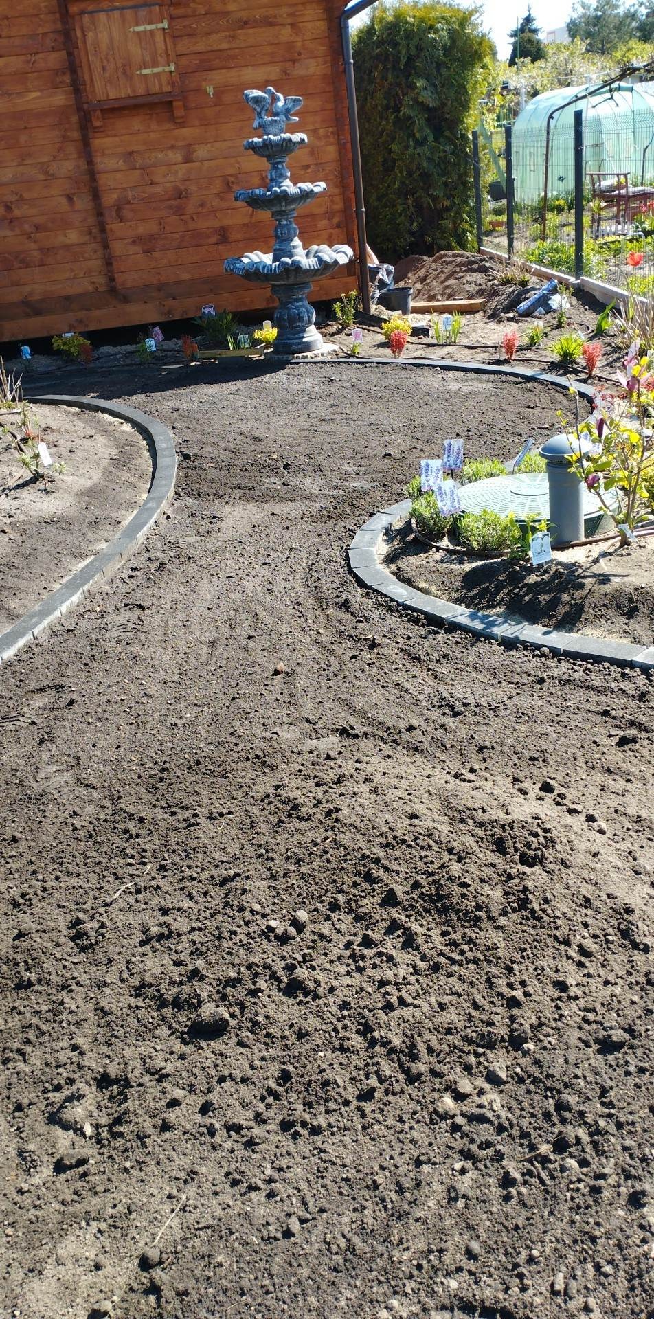 Przygotowany teren pod trawnik z obrzeżami, widoczna fontanna ogrodowa i fragment drewnianej altany w tle, w oddali szklarnia.