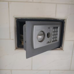 LOCKSTAR Pogotowie zamkowe Artur Gwiazda - Otwarty sejf z panelem numerycznym, wbudowany w ścianę wyłożoną białymi płytkami, widoczne pliki banknotów w środku.