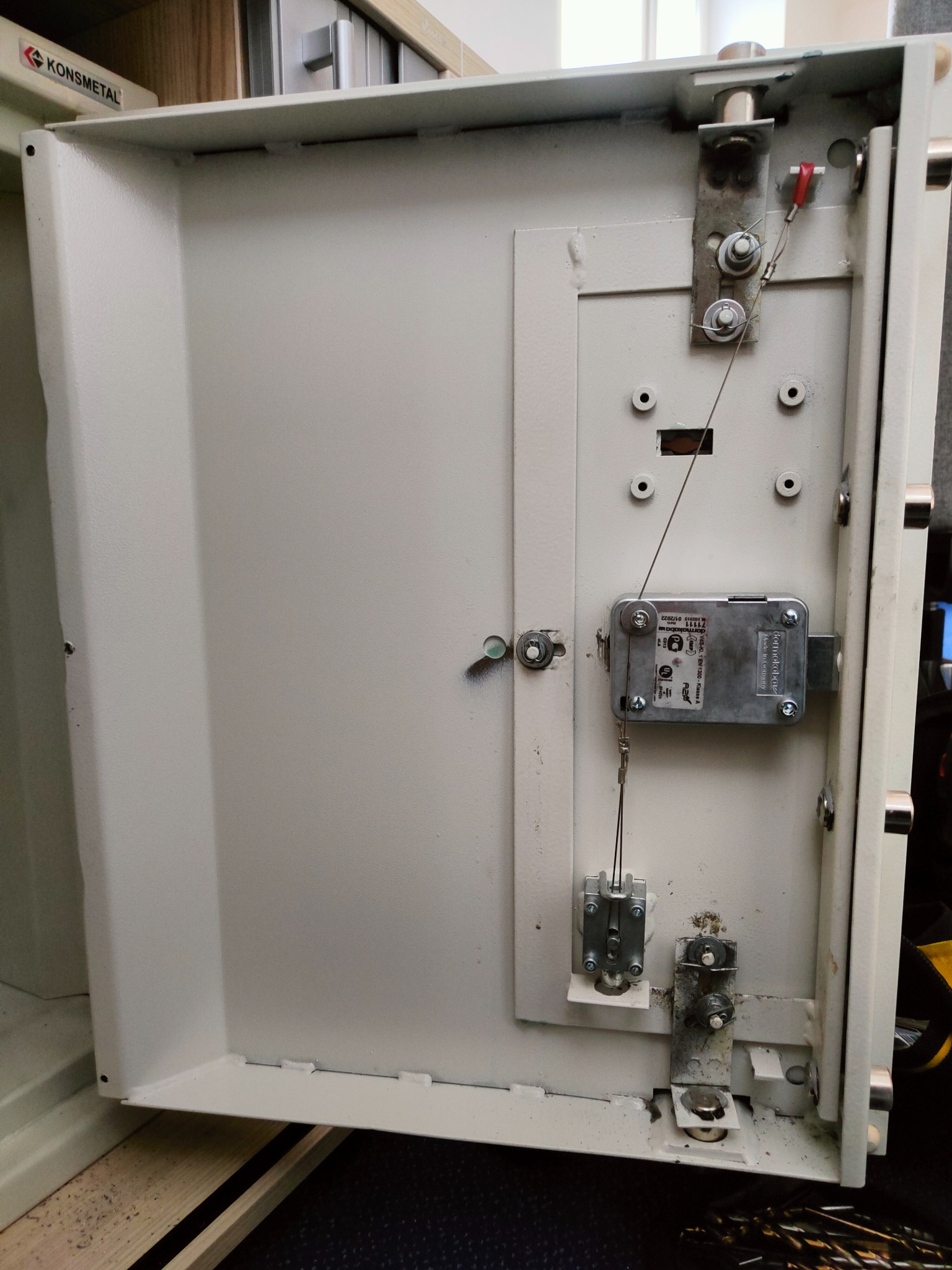 Wnętrze otwartych, metalowych drzwi sejfu marki Konsmetal z widocznym mechanizmem zamka Dormakaba, z elementami mocującymi i stalowymi linkami.