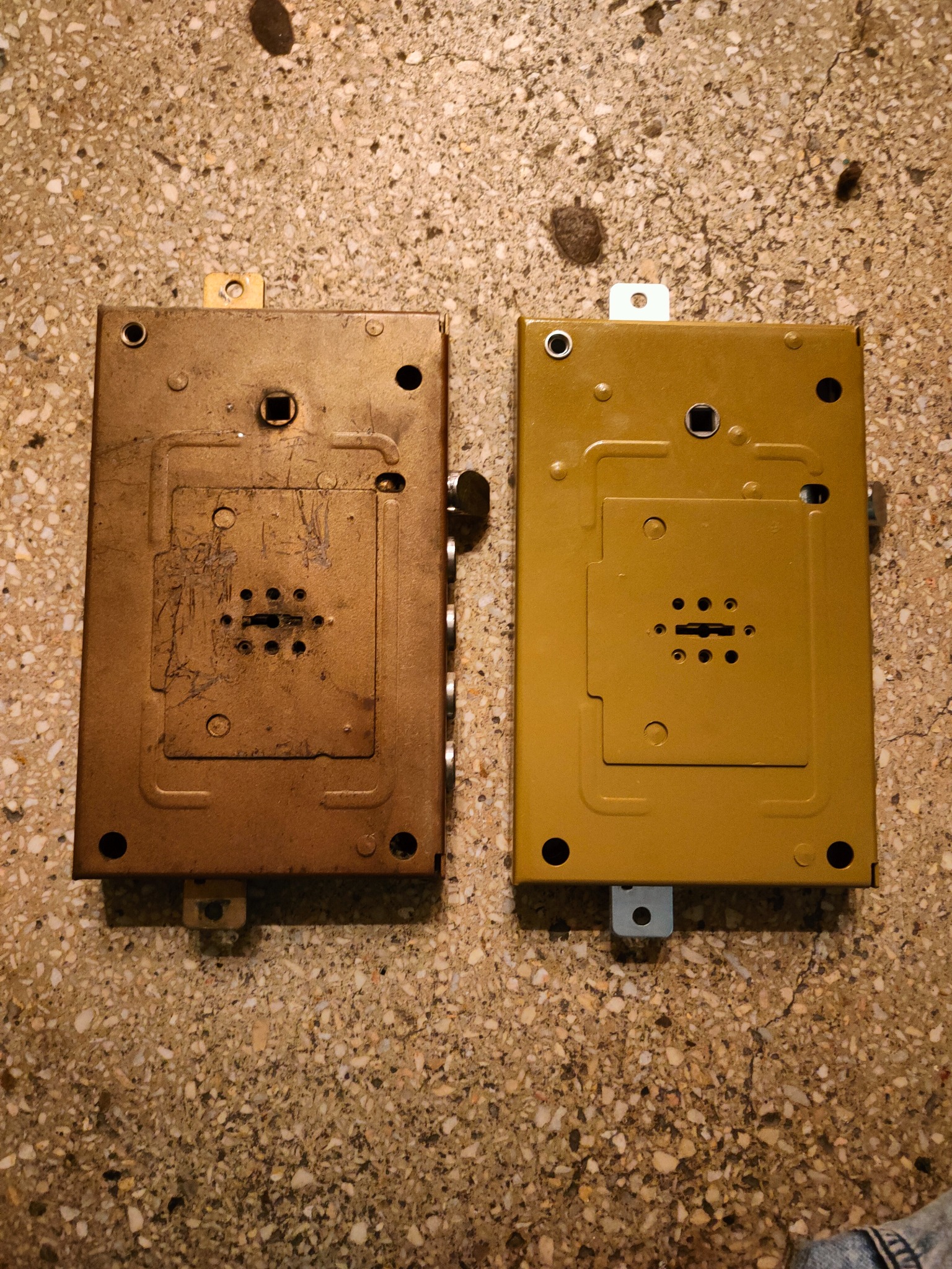 Dwa zamki do drzwi: stary, mosiężny z widocznymi śladami użytkowania i nowy, pomalowany na złoto, leżące na szorstkiej, kamiennej posadzce.