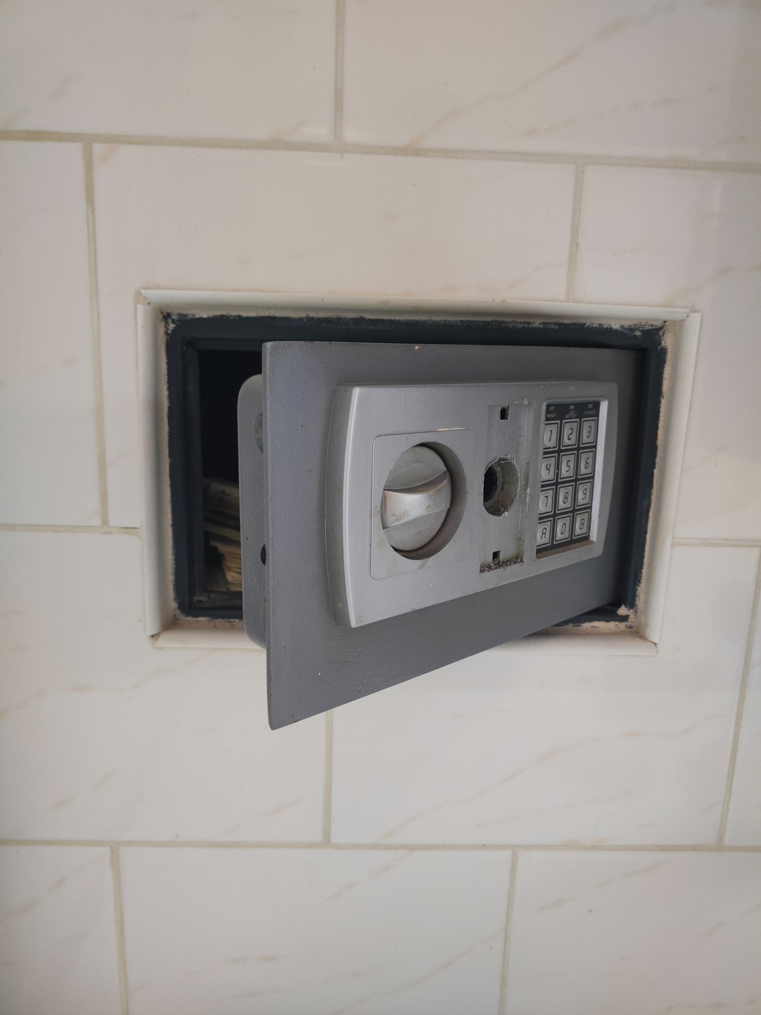Otwarty sejf z panelem numerycznym, wbudowany w ścianę wyłożoną białymi płytkami, widoczne pliki banknotów w środku.