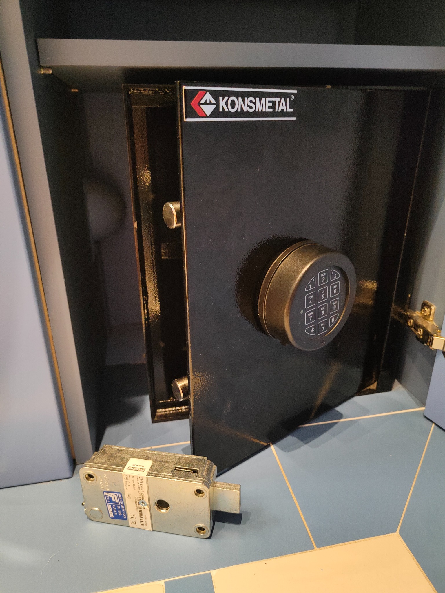 Otwarty sejf firmy Konsmetal z zamkiem szyfrowym i odłożonym zamkiem mechanicznym na podłodze z płytek.