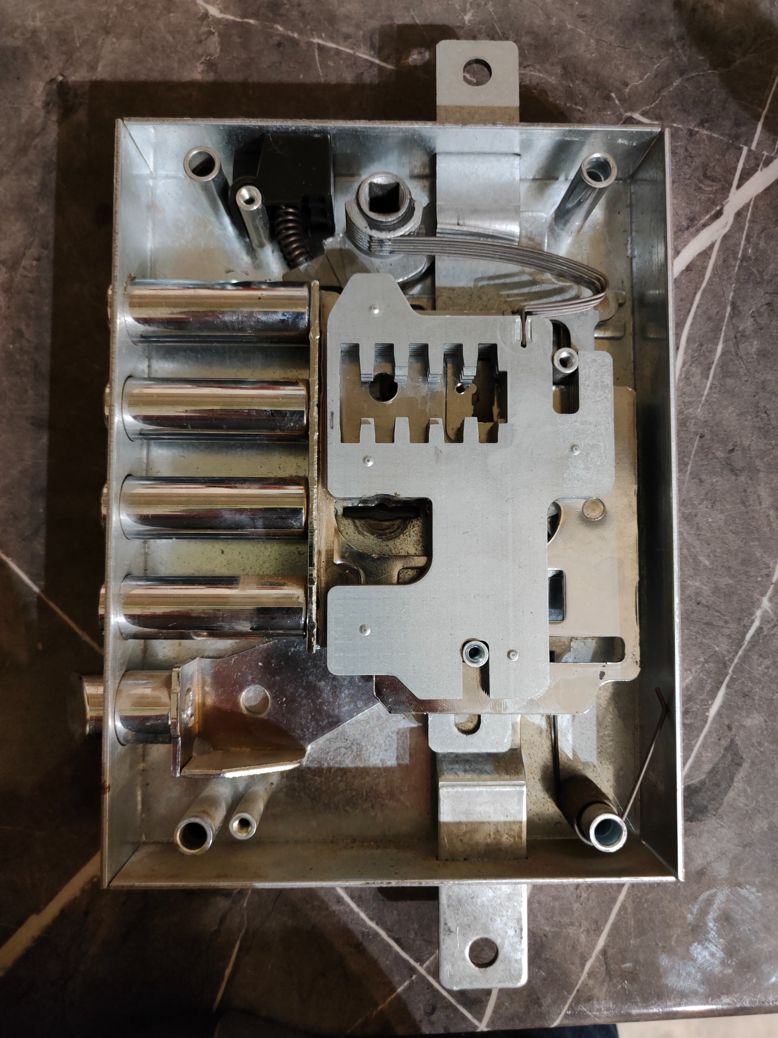 Otwarty mechanizm zamka drzwiowego z widocznymi cylindrami, sprężynami i metalowymi elementami składowymi na tle marmurowej powierzchni.