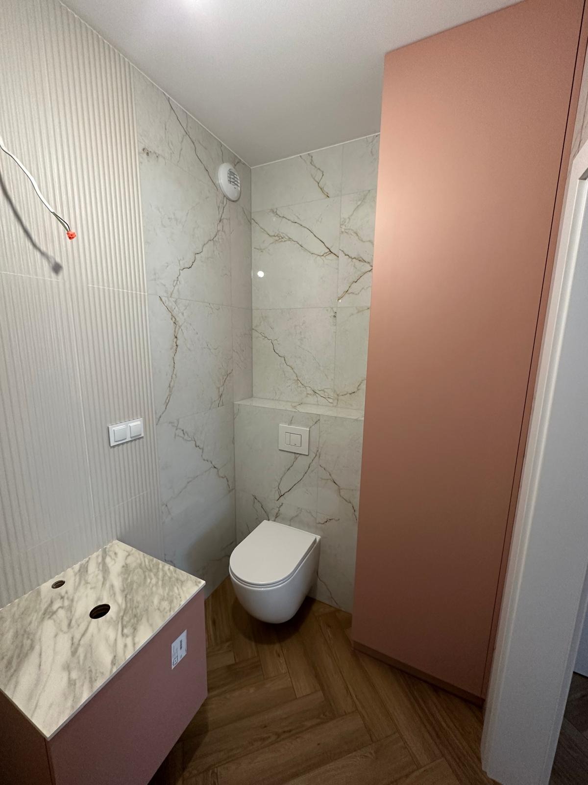 Nowoczesna łazienka z wiszącą toaletą, marmurowymi płytkami, szafką pod umywalkę z blatem imitującym marmur oraz podłogą z drewnopodobnych płytek ułożonych w jodełkę. Widoczne wystające kable...