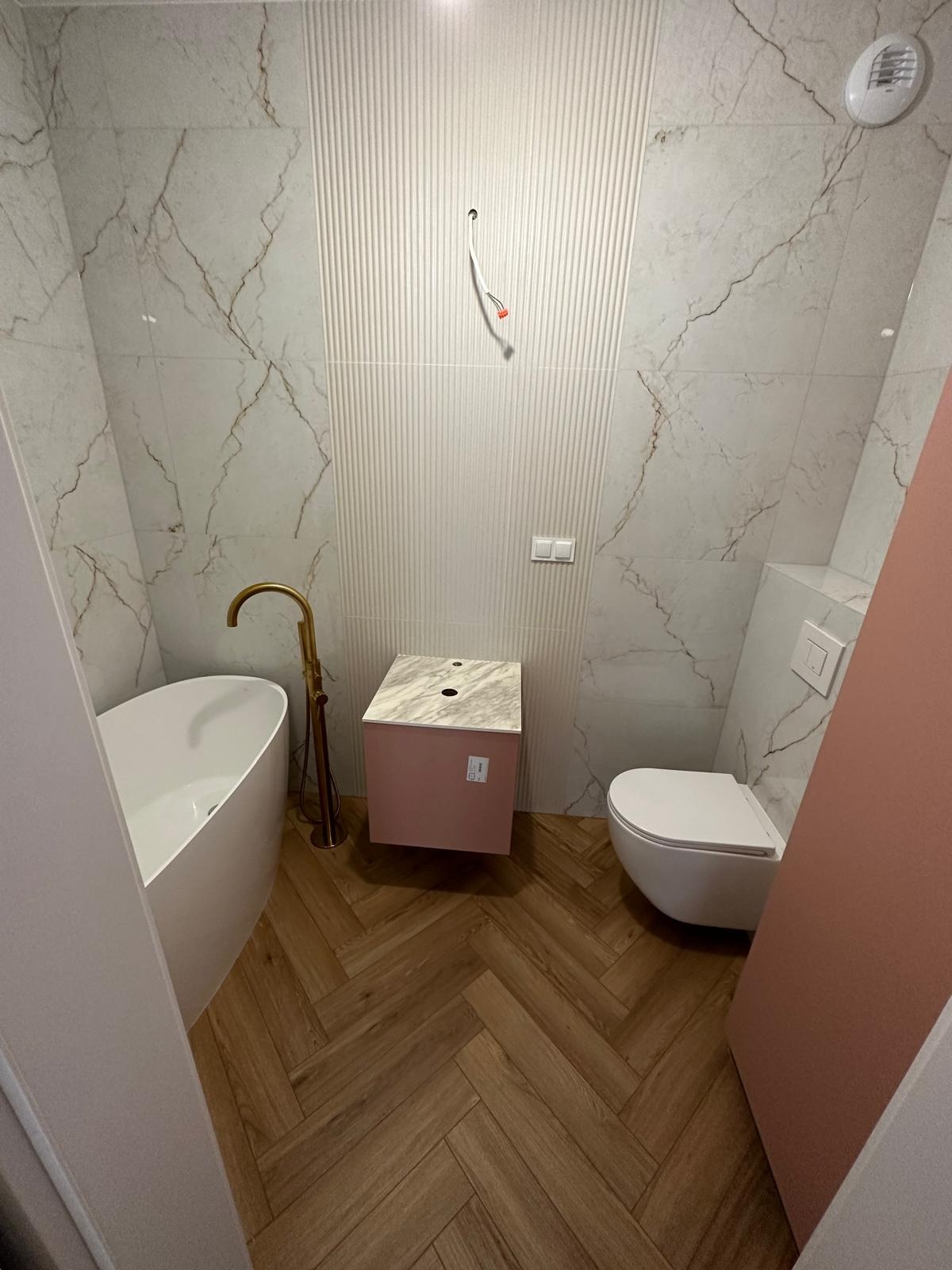 Nowoczesna łazienka w trakcie wykańczania: biała wanna wolnostojąca z złotą baterią, różowa szafka pod umywalkę z marmurowym blatem, podwieszana toaleta, płytki imitujące marmur na ścianach...