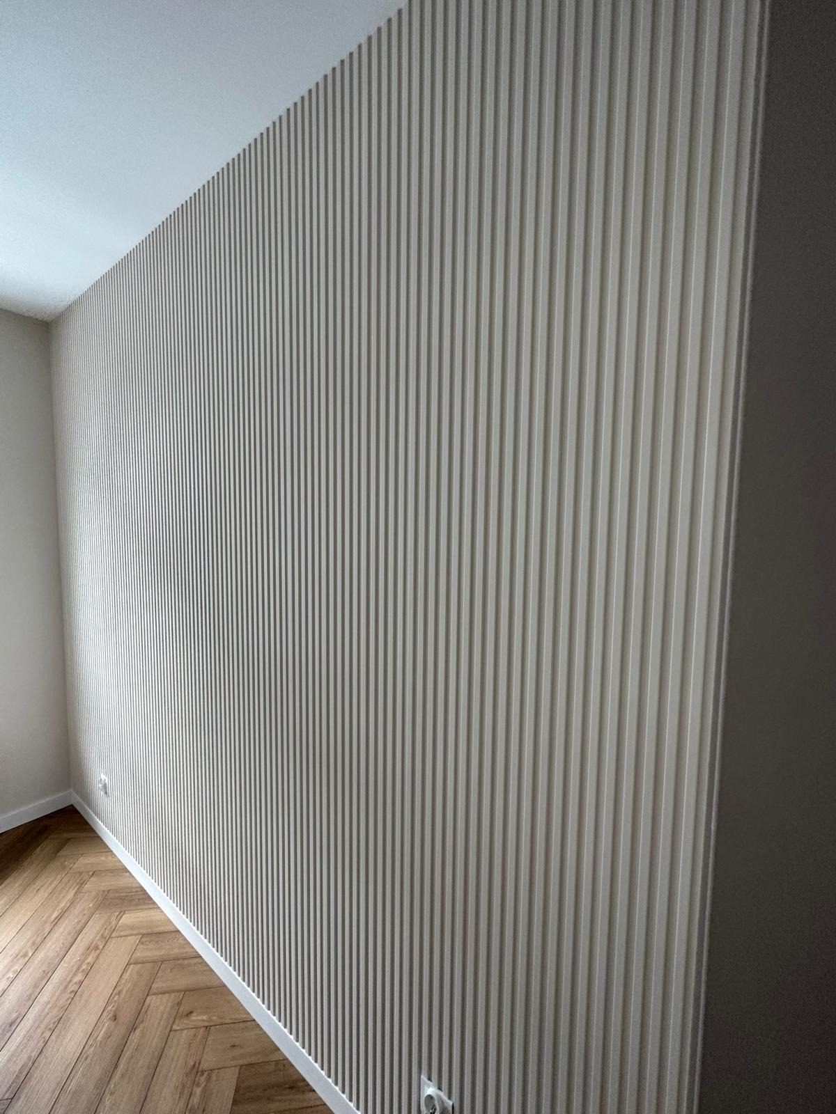 Ściana z panelami ściennymi w pionowe prążki, wykończona listwą przypodłogową, z widocznym gniazdkiem elektrycznym i parkietem ułożonym w jodełkę.