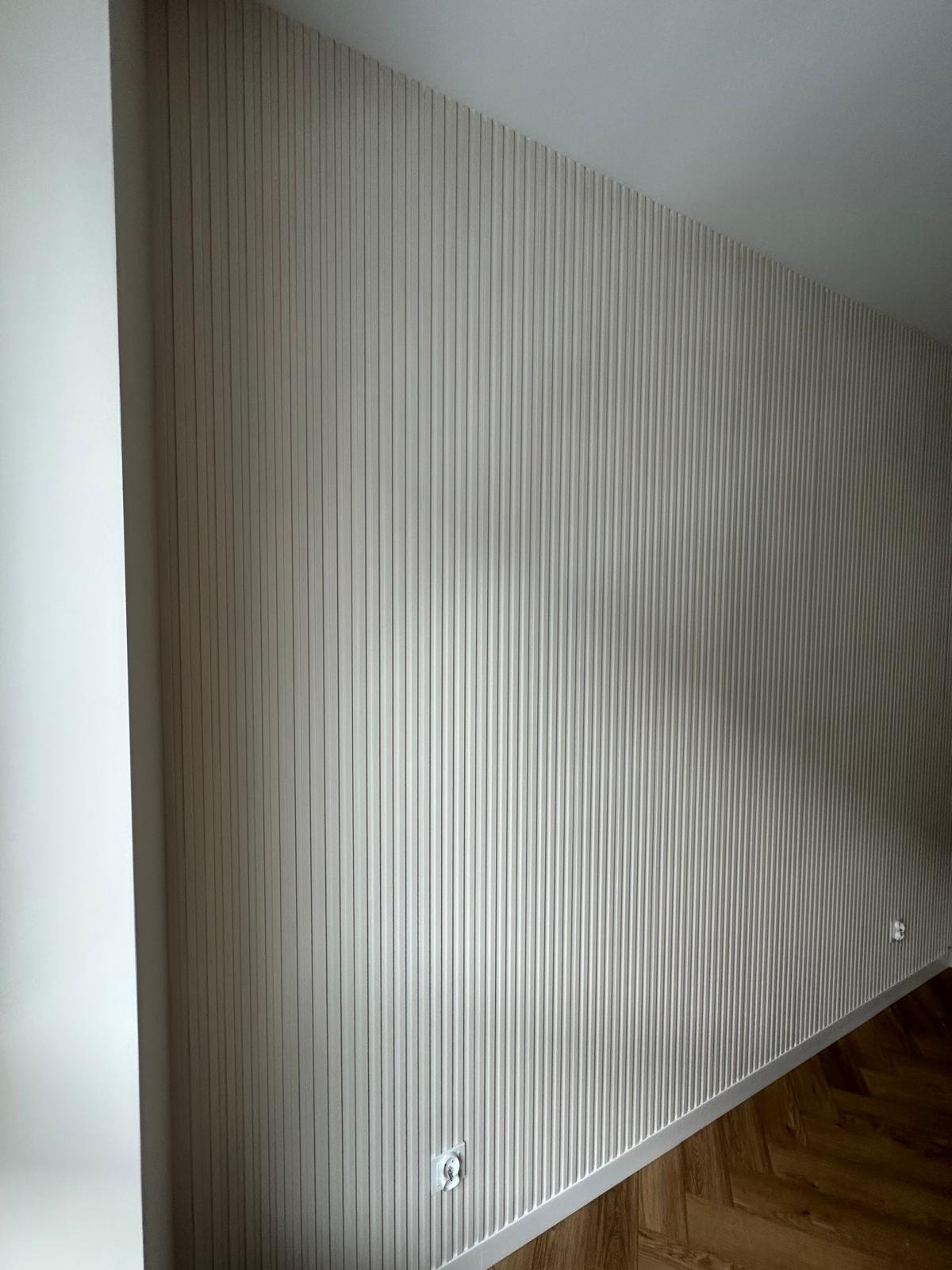 Ściana z dekoracyjnymi panelami pionowymi w odcieniach beżu, widoczna wnęka i fragment parkietu ułożonego w jodełkę, dwa gniazdka elektryczne na ścianie.