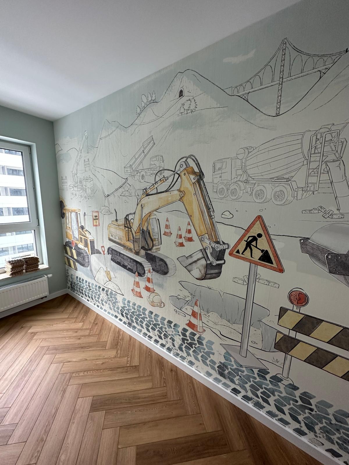 Pokój dziecięcy z tapetą przedstawiającą scenę z budowy: koparki, ciężarówki, znaki ostrzegawcze i krajobraz górski w stylu rysunkowym, uzupełniony parkietem ułożonym w jodełkę.