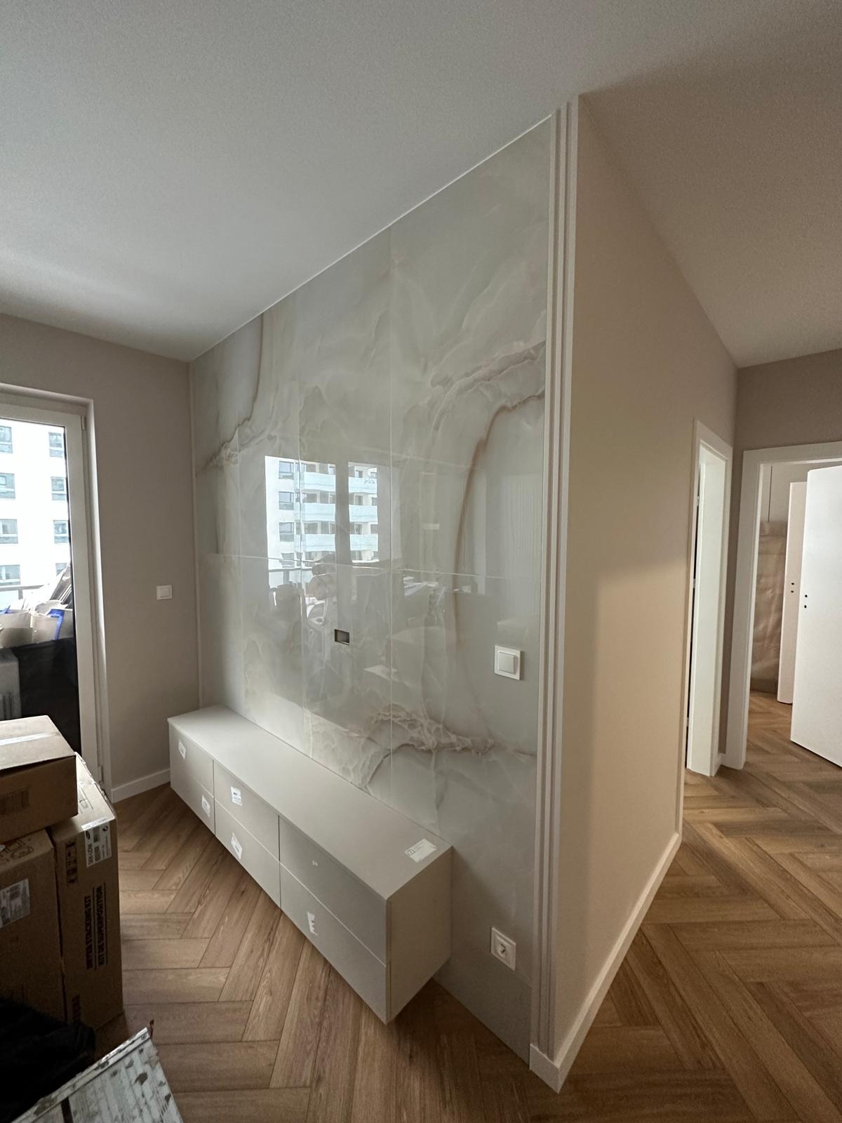 Salon po metamorfozie: ściana wyłożona płytami imitującymi marmur z subtelnym żyłowaniem, dopasowana szafka RTV z szufladami, parkiet ułożony we wzór jodełki francuskiej, neutralne ściany...