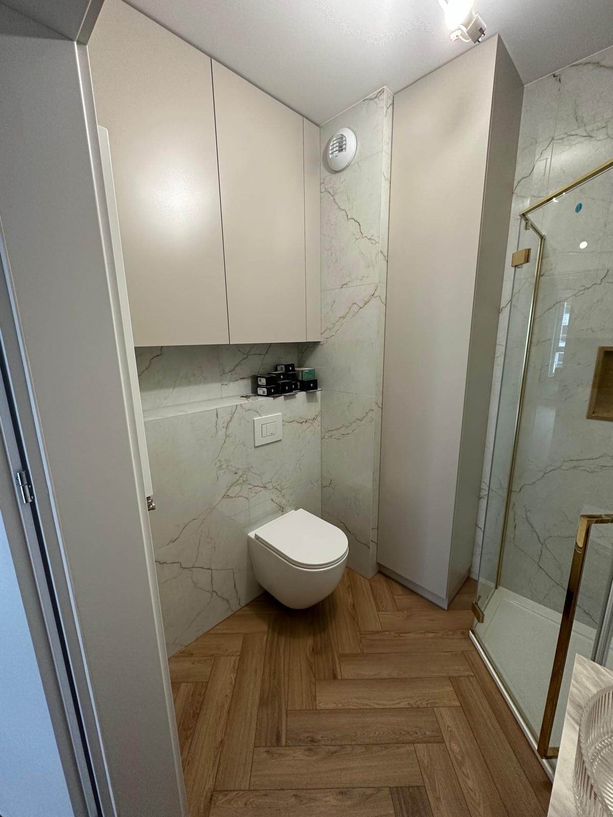 Nowoczesna łazienka z podwieszaną toaletą, kabiną prysznicową ze złotymi detalami, szafkami w kolorze beżowym oraz podłogą z płytek imitujących drewno ułożonych w jodełkę.