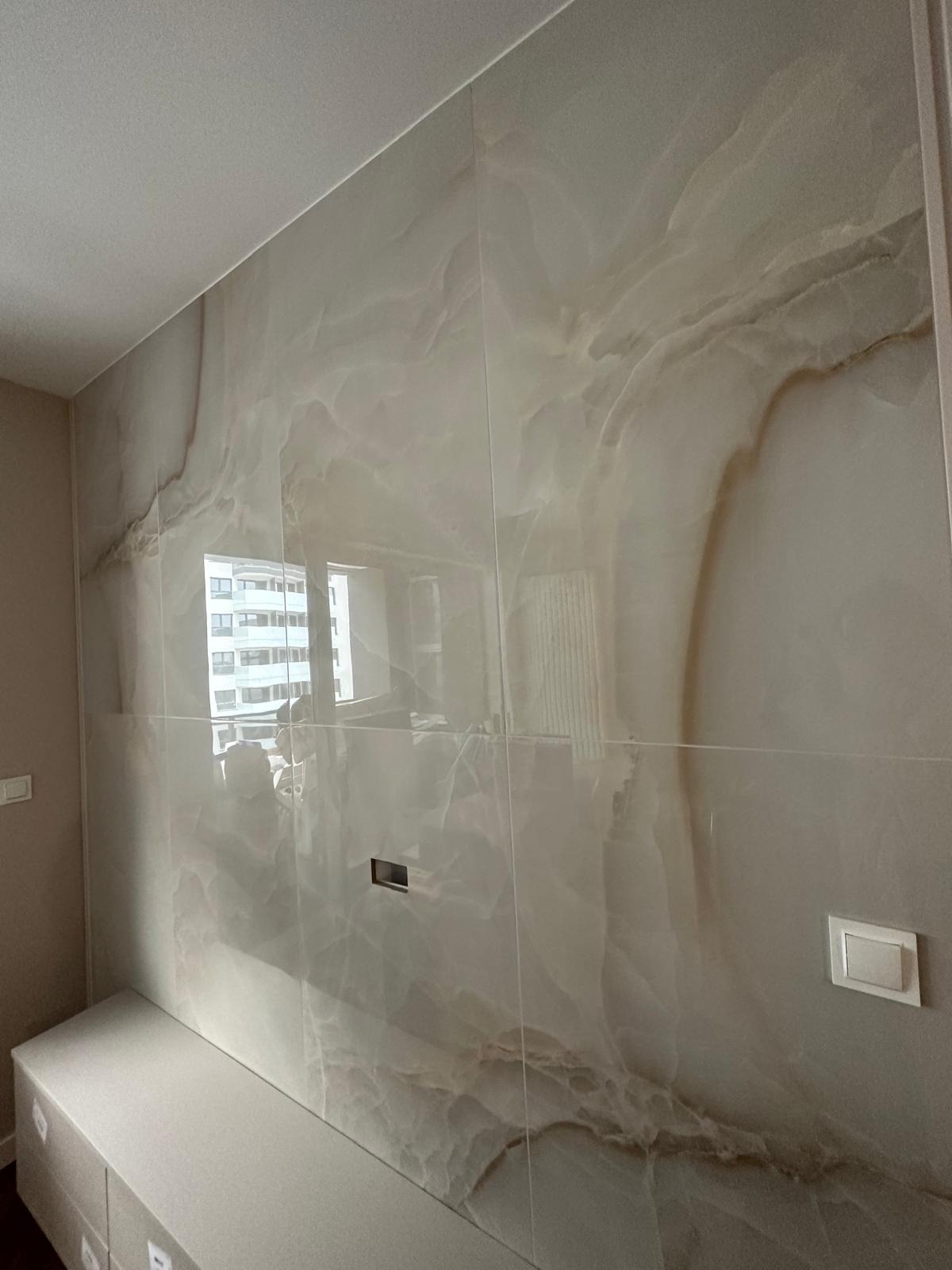 Ściana wyłożona wielkoformatowymi płytami imitującymi marmur, z widocznymi fugami, odbijająca okno budynku naprzeciwko, z zamontowanym panelem sterowania i włącznikiem światła.