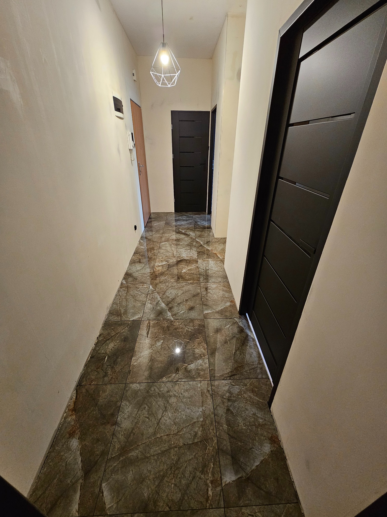 Korytarz w trakcie prac wykończeniowych: ściany przygotowane do malowania, marmurowe płytki podłogowe, nowoczesne czarne drzwi wewnętrzne, minimalistyczna lampa wisząca.
