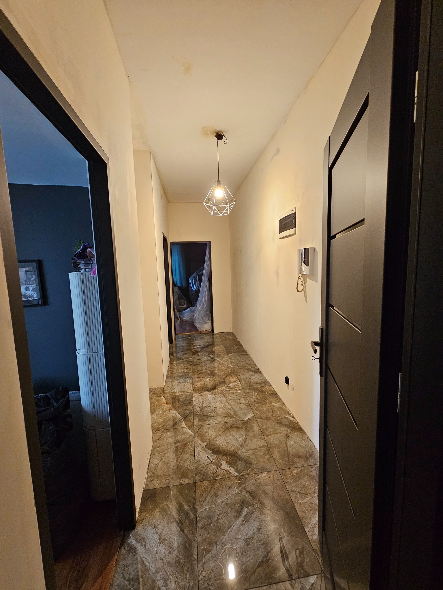 Korytarz w trakcie remontu z położoną marmurkową podłogą, ściany przygotowane do malowania, widoczny interkom i czarne drzwi z poziomymi frezami.