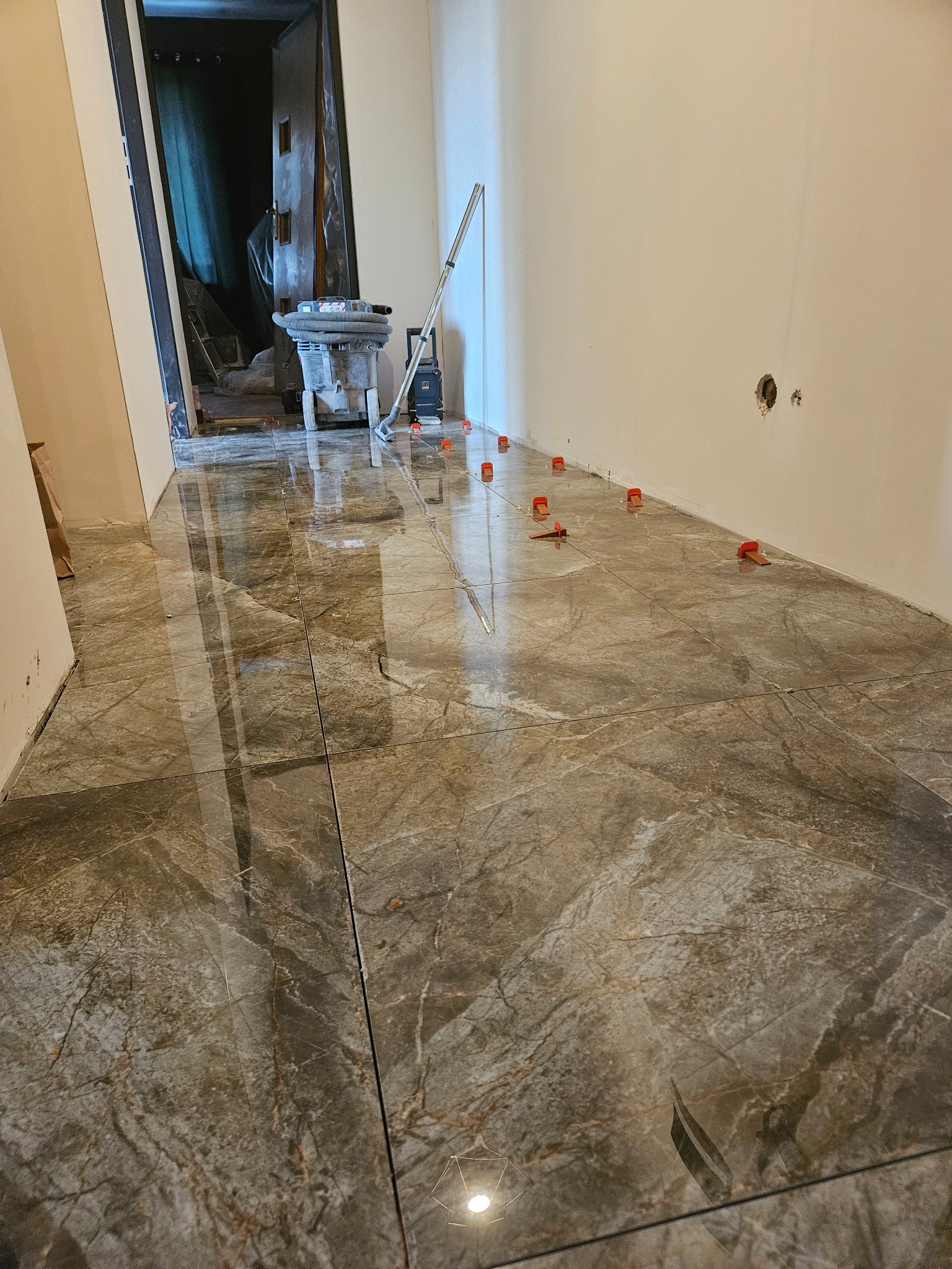 Układanie płytek podłogowych imitujących marmur z użyciem klipsów poziomujących. Widoczny odkurzacz przemysłowy i narzędzia w tle.