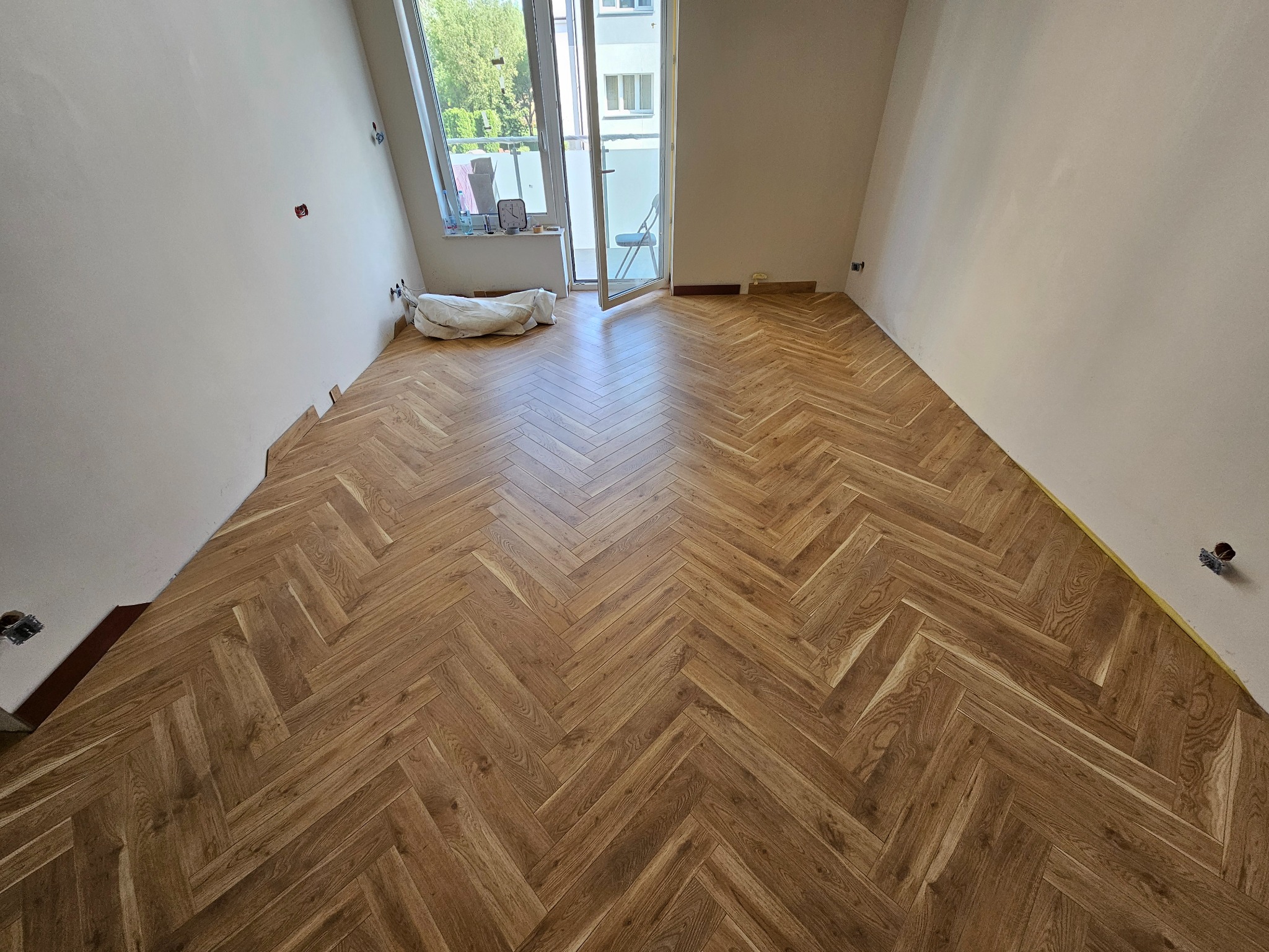 Ukończona podłoga z paneli ułożonych w jodełkę w jasnym pokoju z balkonem, widoczne niedokończone listwy przypodłogowe i zakryte materiałem budowlanym okno.