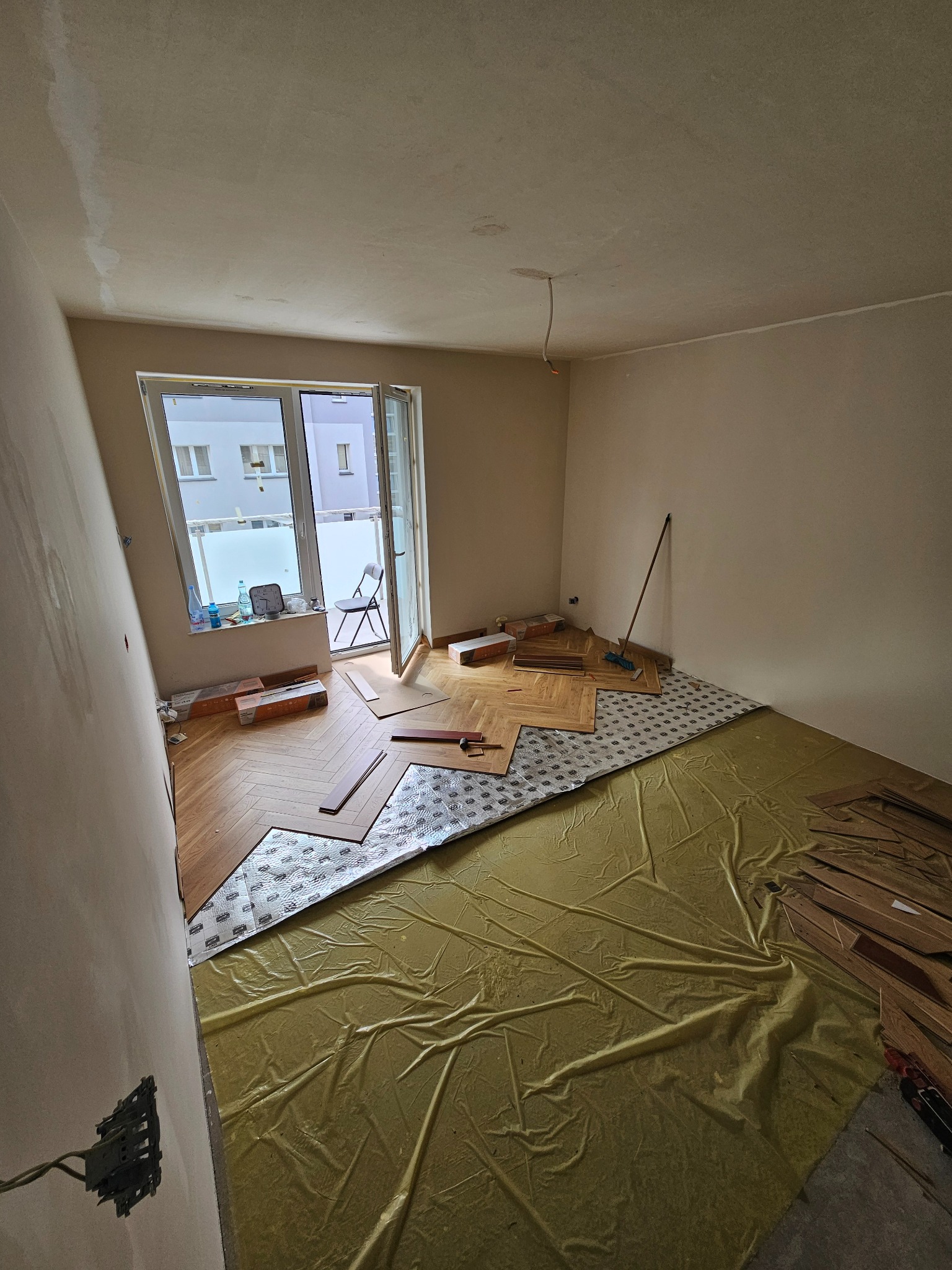 Montaż parkietu w jodełkę: widok na rozłożoną folię, podkład, częściowo ułożony parkiet oraz narzędzia w surowym wnętrzu z balkonem.