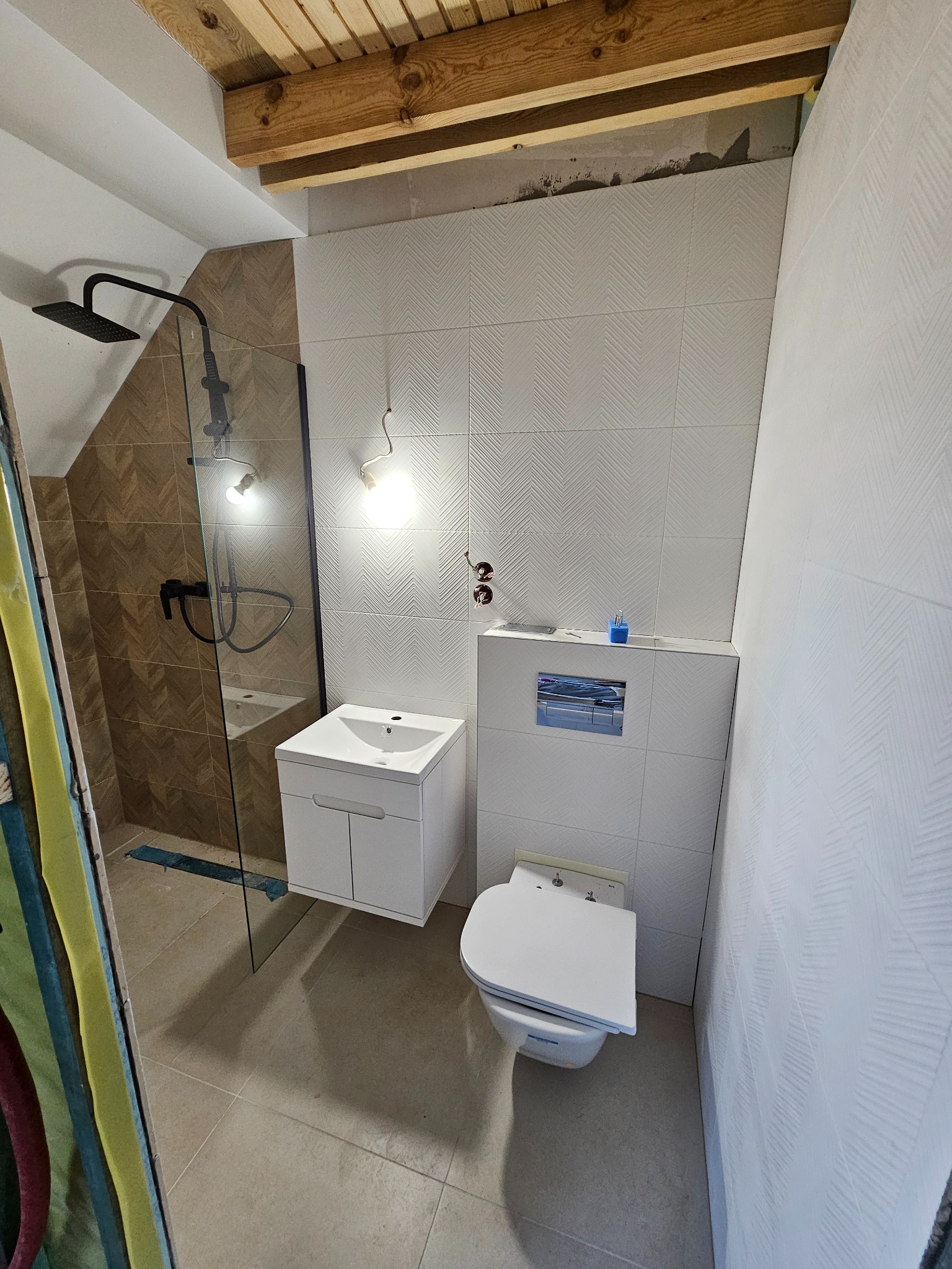 Nowoczesna łazienka w trakcie wykańczania: kabina prysznicowa z czarną armaturą, umywalka z szafką, toaleta podwieszana, białe płytki z delikatnym wzorem jodełki na ścianach, drewniane belki stropowe.