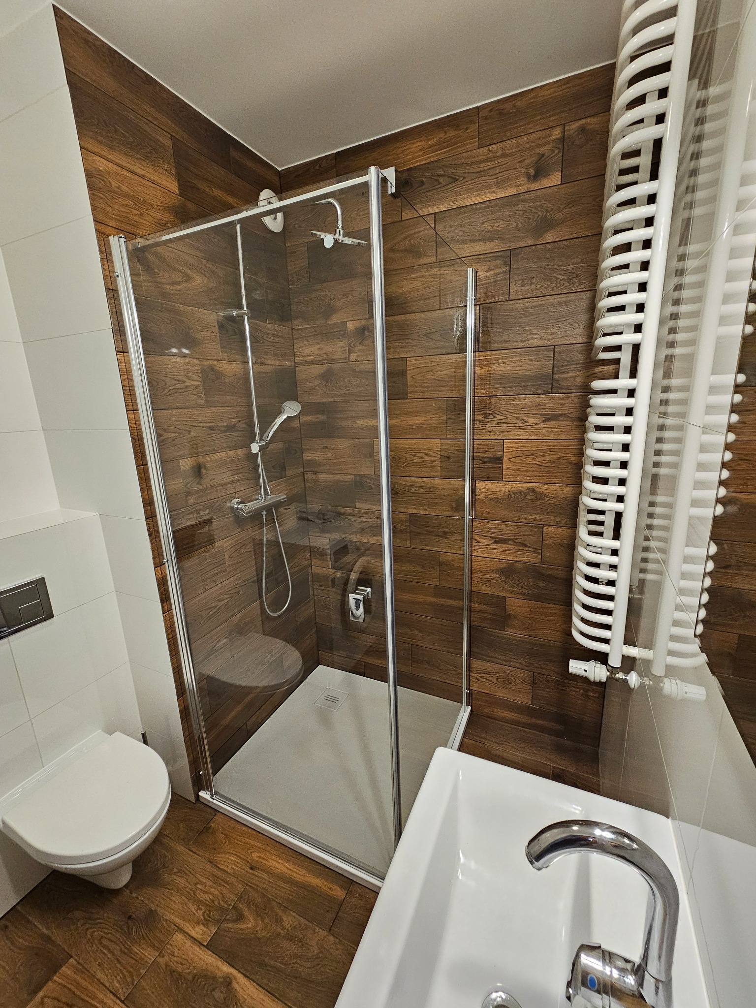 Nowoczesna łazienka z kabiną prysznicową i umywalką, wykończona płytkami imitującymi drewno na ścianach i podłodze, z białymi elementami sanitarnymi i grzejnikiem drabinkowym.