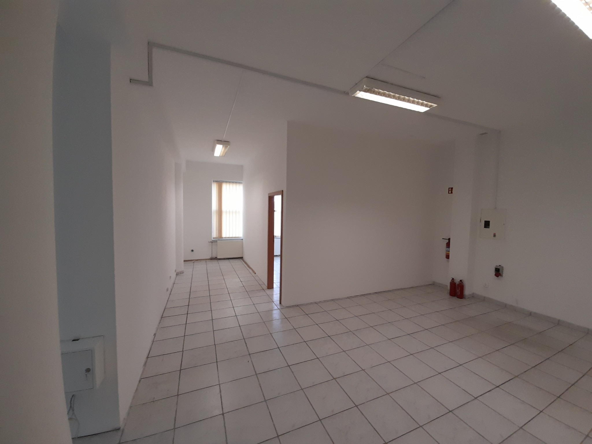 Puste, jasne pomieszczenie z białymi ścianami i podłogą wyłożoną kwadratowymi płytkami, widoczne grzejniki, gaśnice i oświetlenie sufitowe, przygotowane do wykończenia.