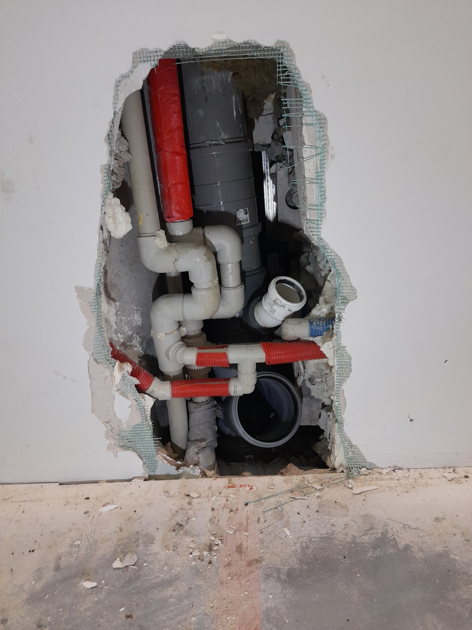 Instalacja wodno-kanalizacyjna w trakcie montażu, widoczne rury z tworzywa sztucznego w kolorach szarym, białym i czerwonym, wystające ze ściany z naruszoną strukturą, zabezpieczoną siatką.