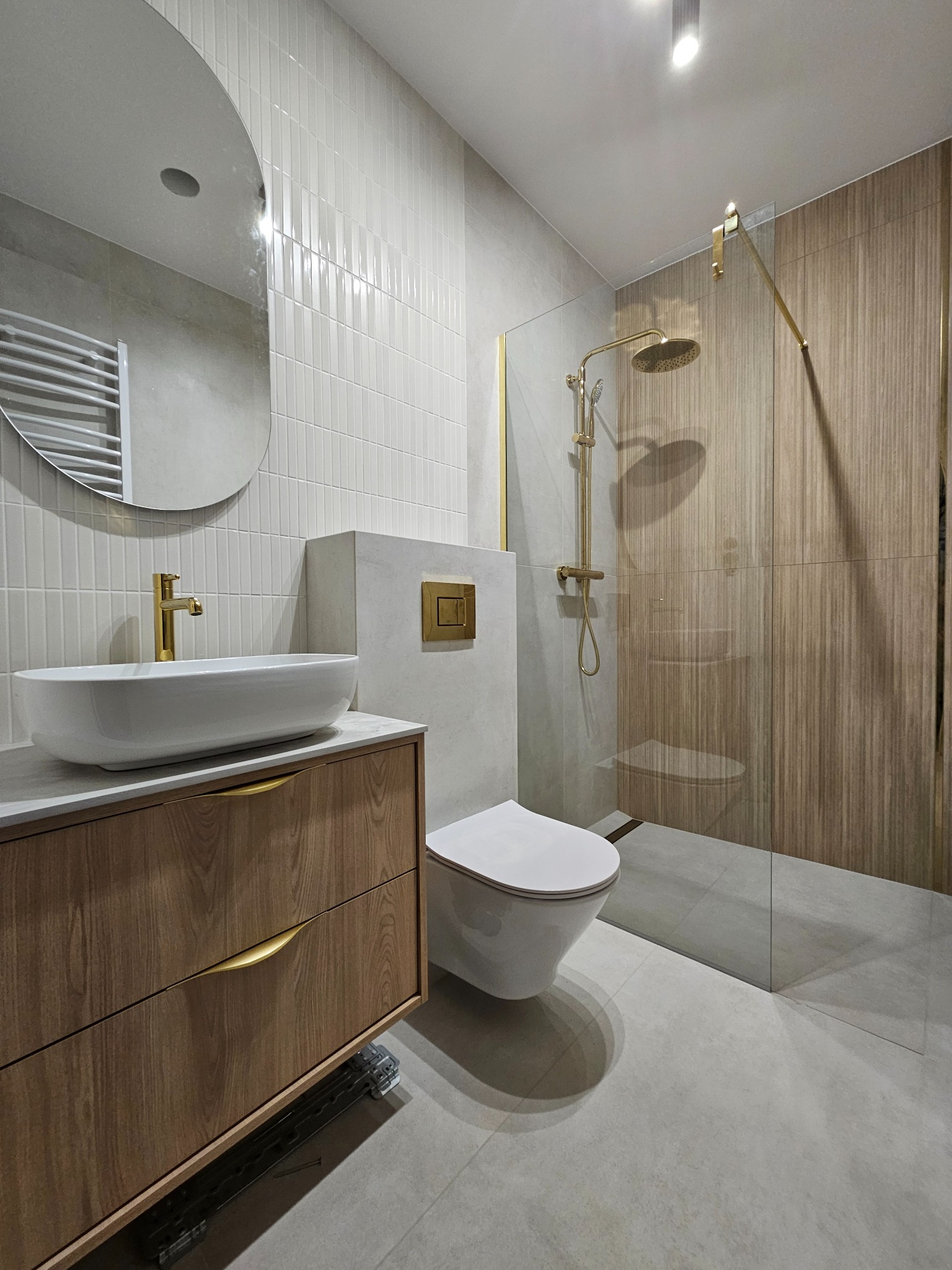 Nowoczesna łazienka z drewnianymi akcentami: szafka pod umywalką, kabina prysznicowa ze złotymi detalami, owalne lustro i płytki w odcieniach bieli i szarości.