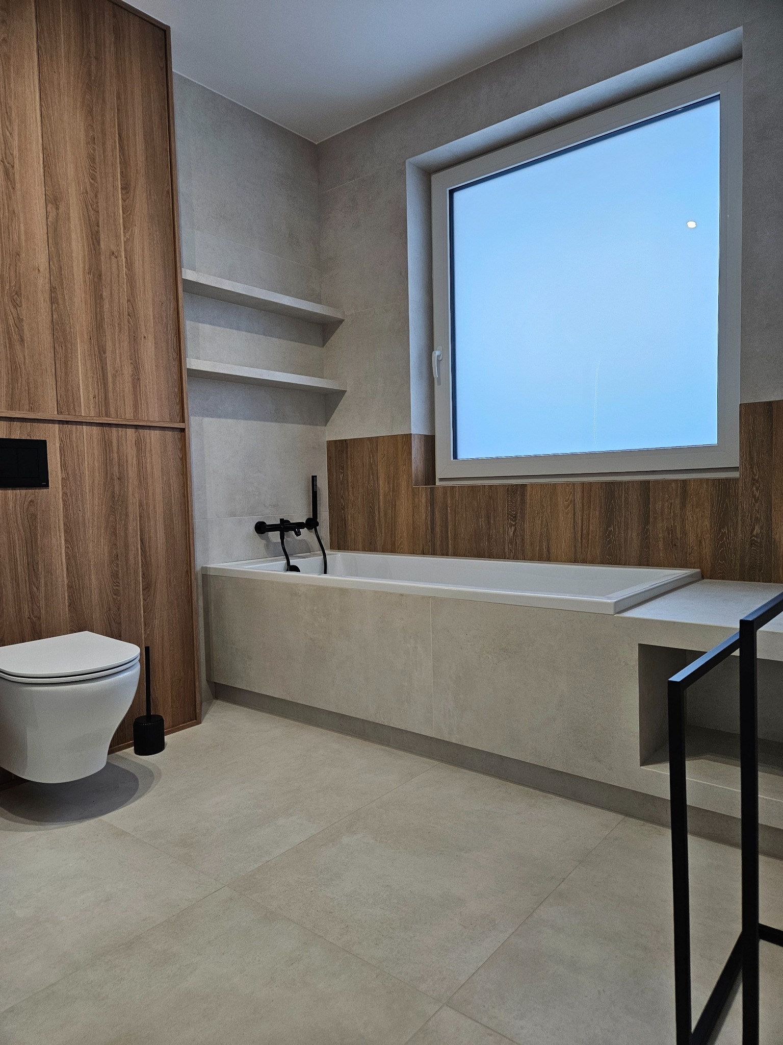 Nowoczesna łazienka z drewnianymi akcentami: zabudowa, półki, obudowa wanny. Minimalistyczna armatura w czerni, jasne płytki podłogowe i ścienne. Duże okno.