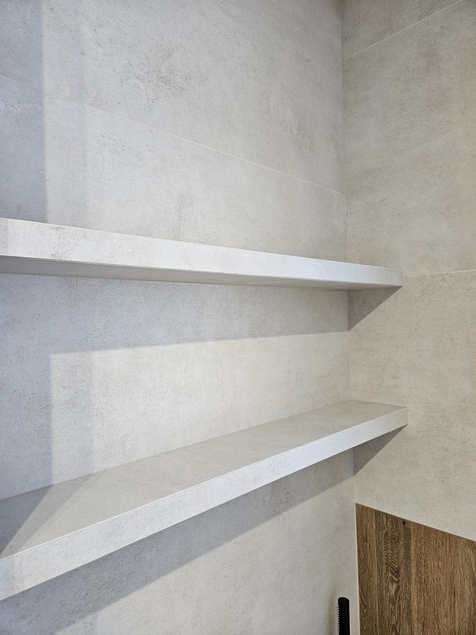 Minimalistyczna łazienka z dwoma półkami z jasnego kamienia, dopasowanymi do płytek ściennych. U dołu widoczny fragment drewnopodobnej okładziny.
