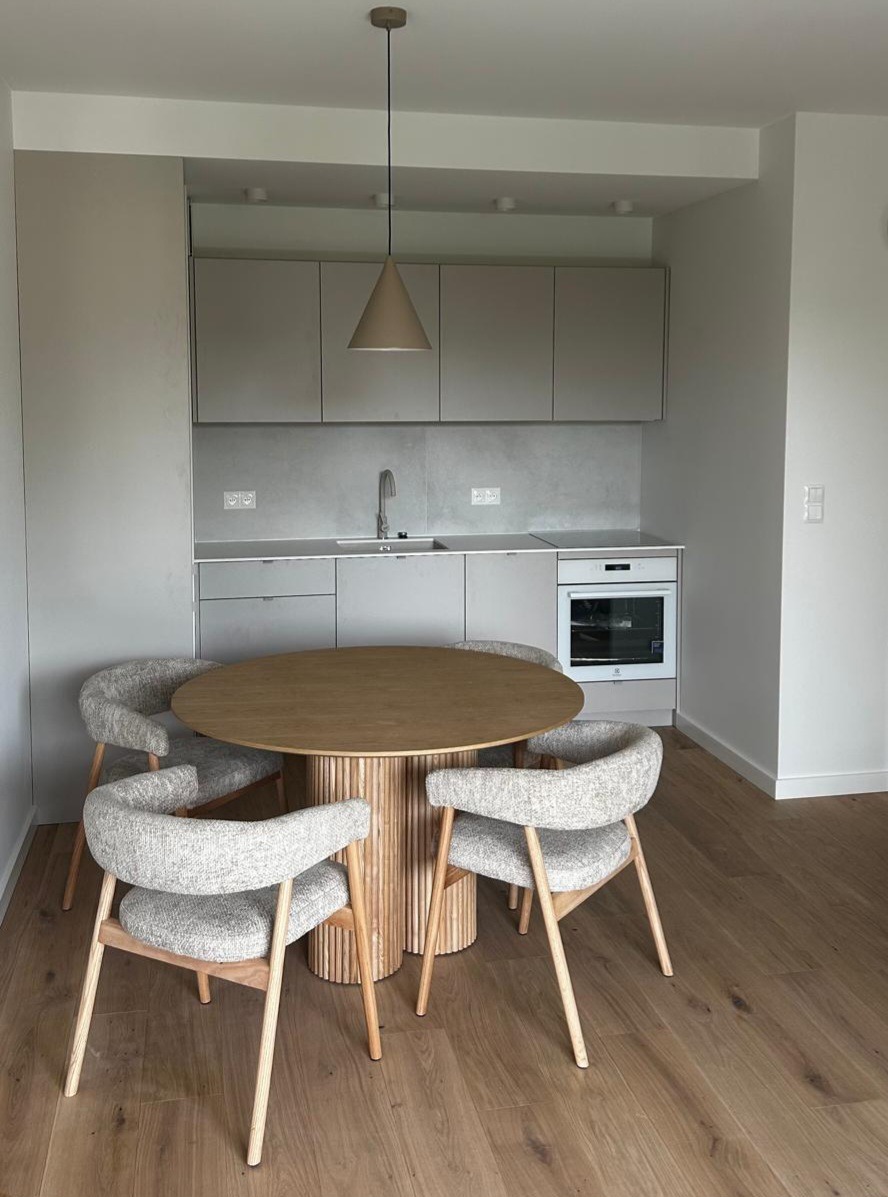 Jasna kuchnia z okrągłym stołem i czterema krzesłami. Minimalistyczny design, drewniana podłoga, neutralna kolorystyka. Widok na zabudowę kuchenną z piekarnikiem.