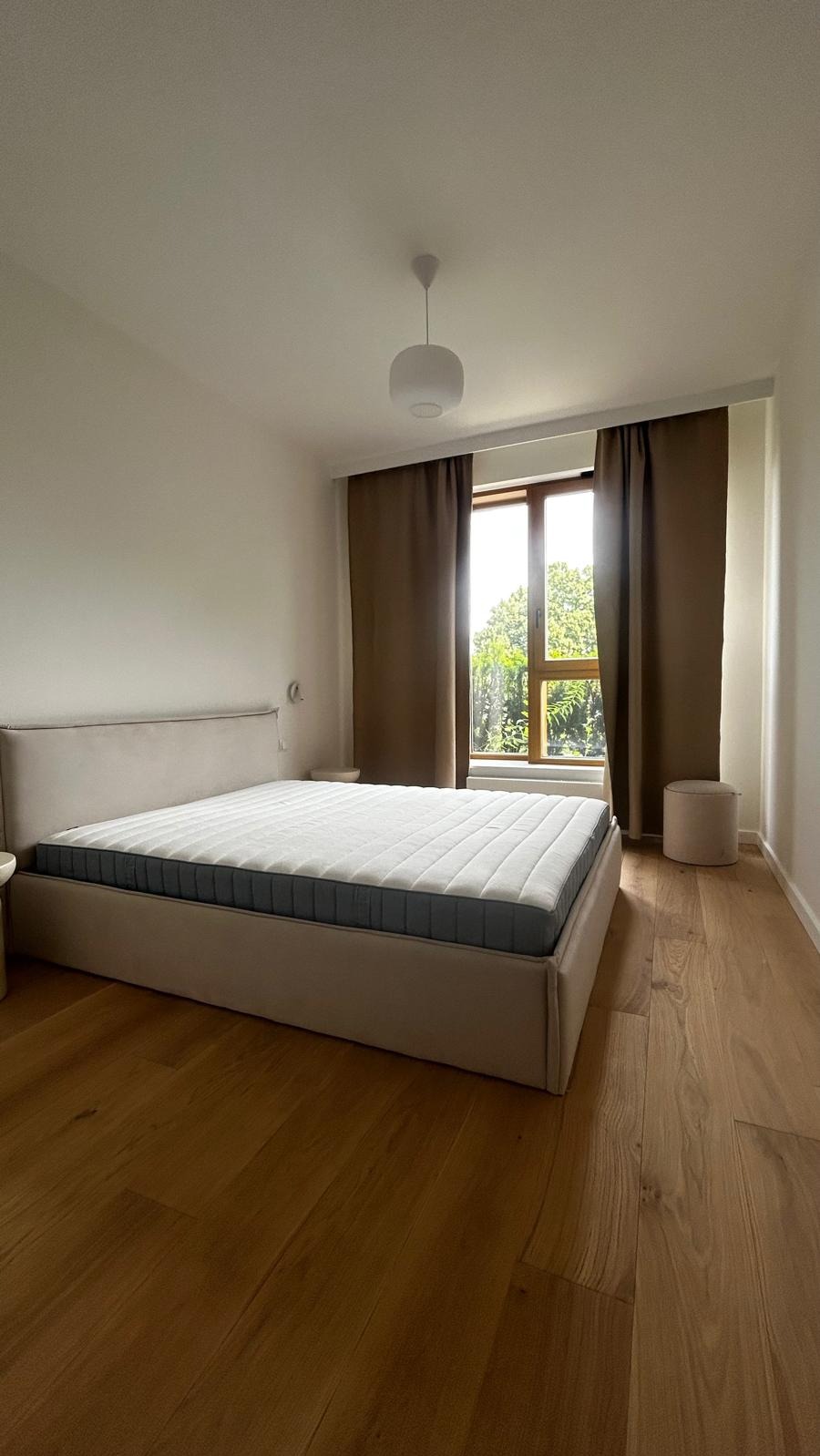 Jasna sypialnia z drewnianą podłogą: beżowe łóżko z materacem, brązowe zasłony przy oknie, okrągły puf. Minimalistyczny design, naturalne światło.