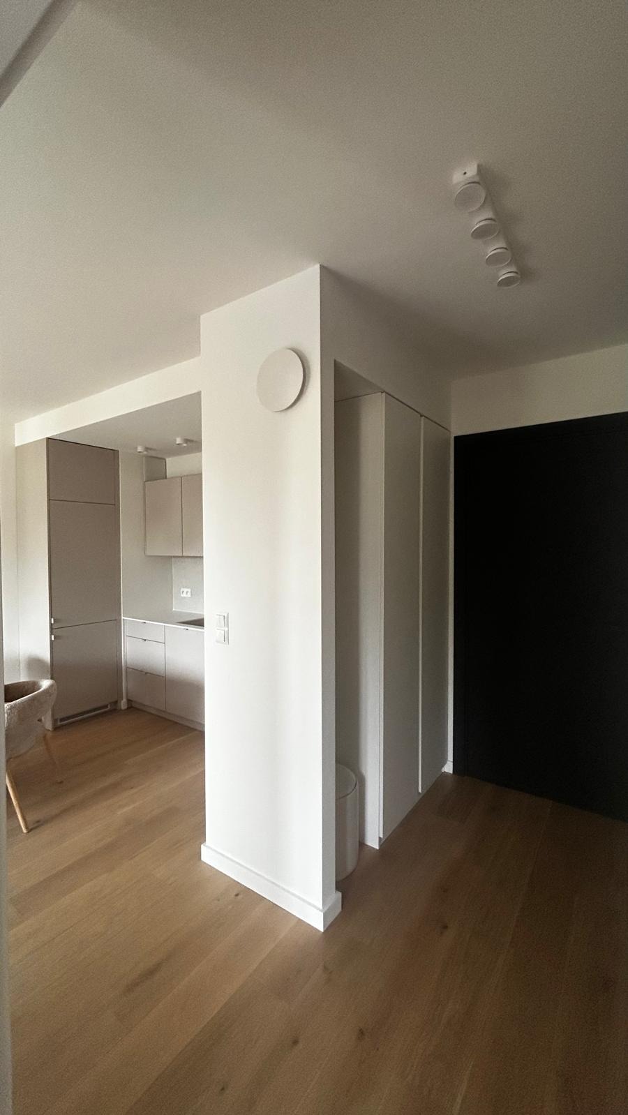 Jasne, minimalistyczne wnętrze z drewnianą podłogą, widok na kuchnię i czarne drzwi. Okrągły, biały element dekoracyjny na ścianie. Oświetlenie sufitowe.