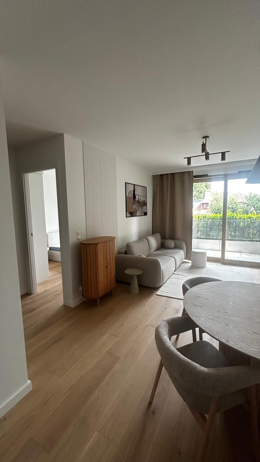 Jasne, przestronne wnętrze mieszkania z drewnianą podłogą, beżową sofą i okrągłym stołem. Widoczna szafka z lamelami i dekoracyjna ściana z pionowymi frezami.