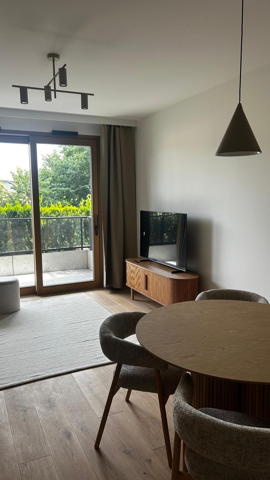 Jasne wnętrze: drewniana podłoga, okrągły stół z krzesłami, szafka RTV z telewizorem, wyjście na balkon z zielenią. Minimalistyczny design, naturalne światło.