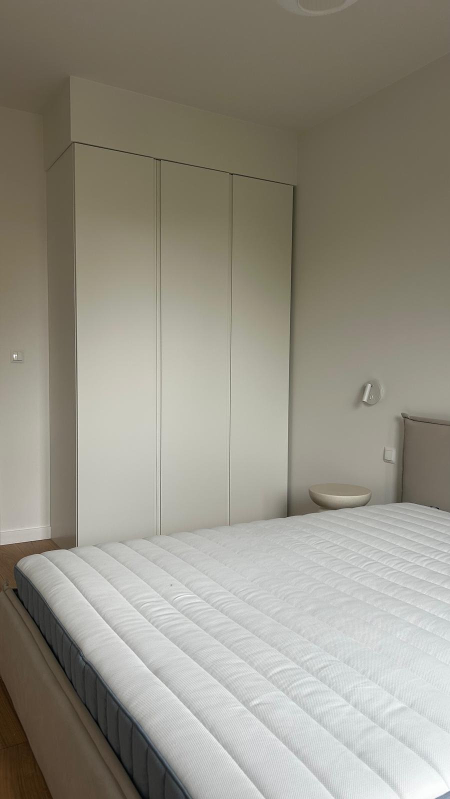 Minimalistyczna sypialnia z dużym, białym łóżkiem i zabudowaną szafą w odcieniach szarości. Na ścianie lampka nocna i stolik. Jasne drewno podłogi.