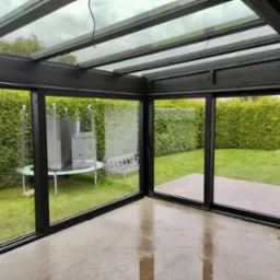 Szklana pergola metalowa z przesuwnymi drzwiami aluminiowymi, widok na ogród z trampoliną i żywopłotem, mokra podłoga odbija światło.