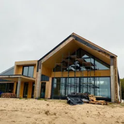 Nowoczesny dom w trakcie budowy z dużymi oknami, widoczne rusztowania i palety z materiałami budowlanymi na piaszczystym terenie.