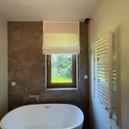 Nowoczesna łazienka z wolnostojącą wanną, brązowymi płytkami, grzejnikiem drabinkowym i oknem z jasną roletą rzymską, przez które widać zieleń ogrodu.