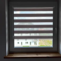 Okno z zamontowaną roletą typu dzień i noc, widok na budynek mieszkalny i fragment zieleni. Roleta w kolorze beżowym z poziomymi pasami.