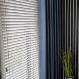 Białe żaluzje poziome przy oknie, obok zasłona w odcieniach niebieskiego, dekoracyjna ściana w czarno-białe pasy oraz roślina doniczkowa na ciemnej podłodze.