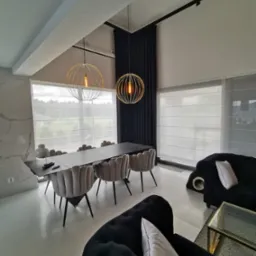 Nowoczesne wnętrze z wysokim sufitem, oświetlone złotymi lampami wiszącymi nad stołem jadalnym z szarymi krzesłami, z widocznymi białymi roletami w oknach i ciemnymi zasłonami oraz czarnymi sofami...