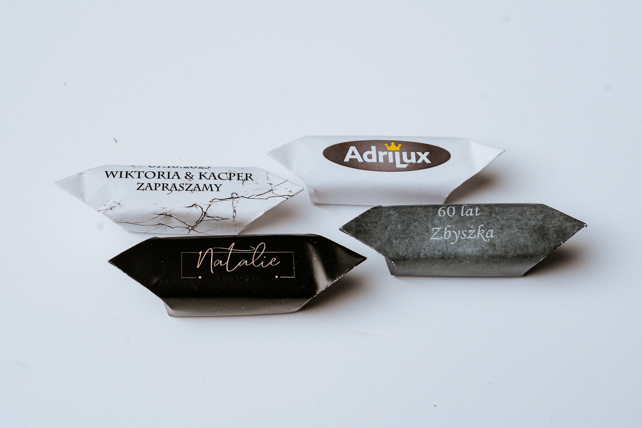 Cztery spersonalizowane cukierki w ozdobnych papierkach: zaproszenie ślubne Wiktoria & Kacper, logo Adrilux z koroną, napis 60 lat Zbyszkowi, oraz logo Natalie Boutique.