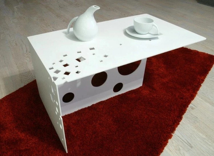 Nowoczesny biały stolik kawowy z geometrycznymi wycięciami i okrągłymi otworami, na czerwonym puszystym dywanie, z białym dzbankiem i filiżanką.