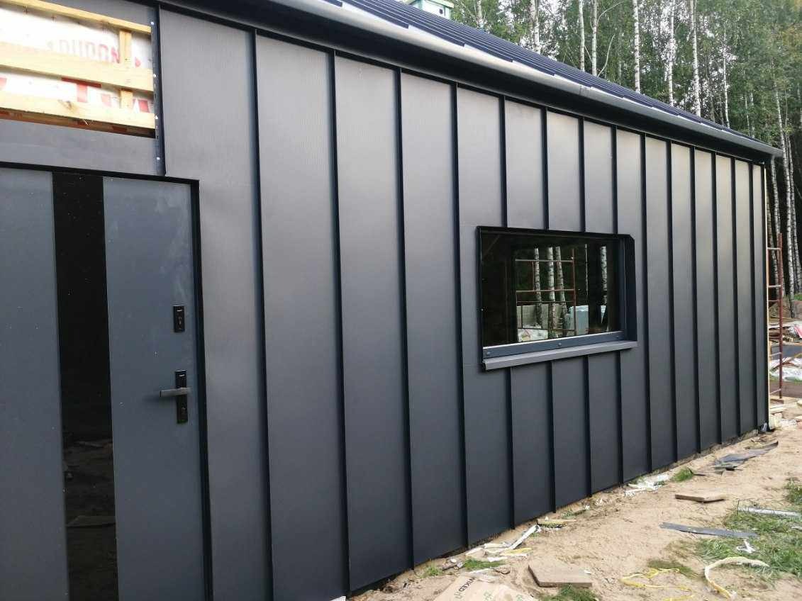 Nowoczesna, szara elewacja budynku z blachy trapezowej, z oknem i drzwiami, na tle drzew.