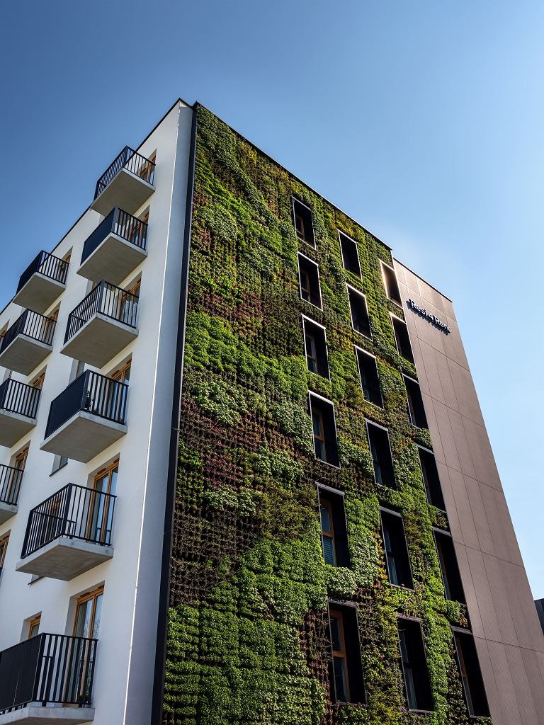 Wielopiętrowy budynek z pionowym ogrodem na elewacji, balkony z czarnymi balustradami i błękitne niebo w tle.