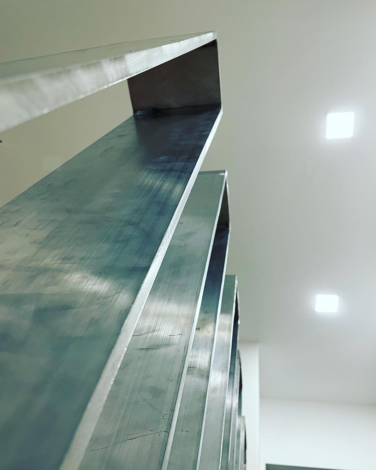 Metalowe elementy konstrukcyjne ułożone jeden za drugim, widok od dołu w kierunku sufitu z kwadratowymi lampami.