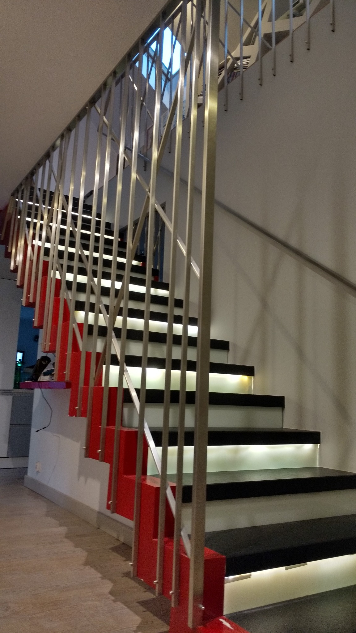 Nowoczesne schody z podświetleniem LED, czarnymi stopniami, czerwonymi wspornikami i metalową balustradą.